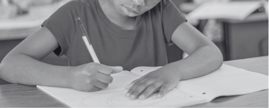 Criança negra escrevendo em um caderno com lápis, sentada à carteira escolar. Outras crianças desfocadas ao fundo também estão escrevendo.