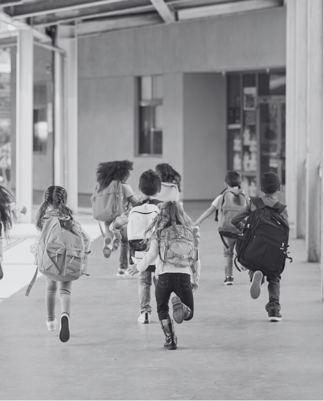Grupo de crianças pequenas correndo por um corredor coberto em direção à entrada de uma escola. Todas carregam mochilas nas costas e estão de costas para a câmera, em clima de entusiasmo.